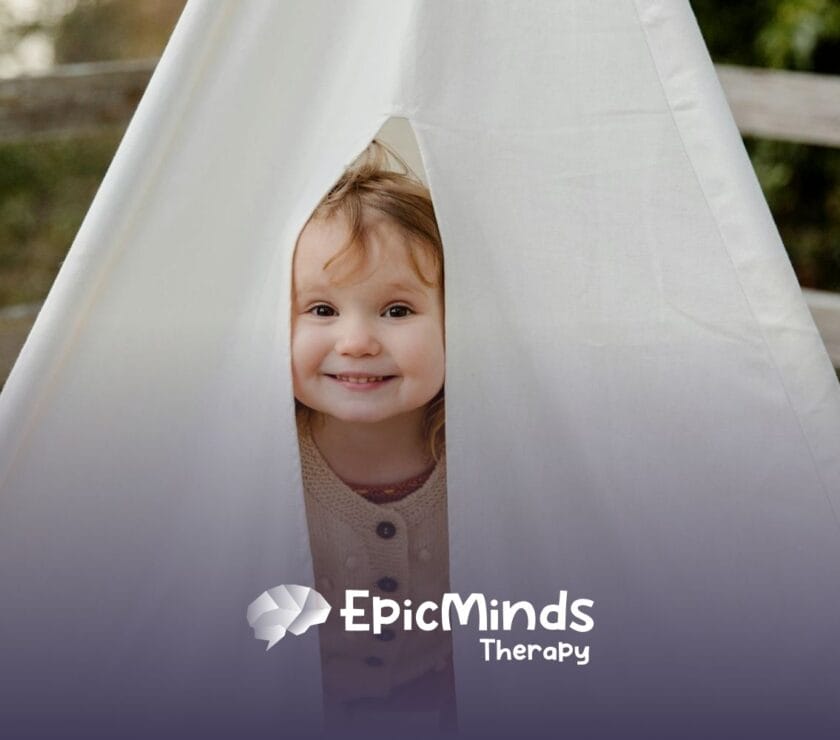 Smiling autistic toddler peeking out from inside a white play tent outdoors during ABA therapy in NC.