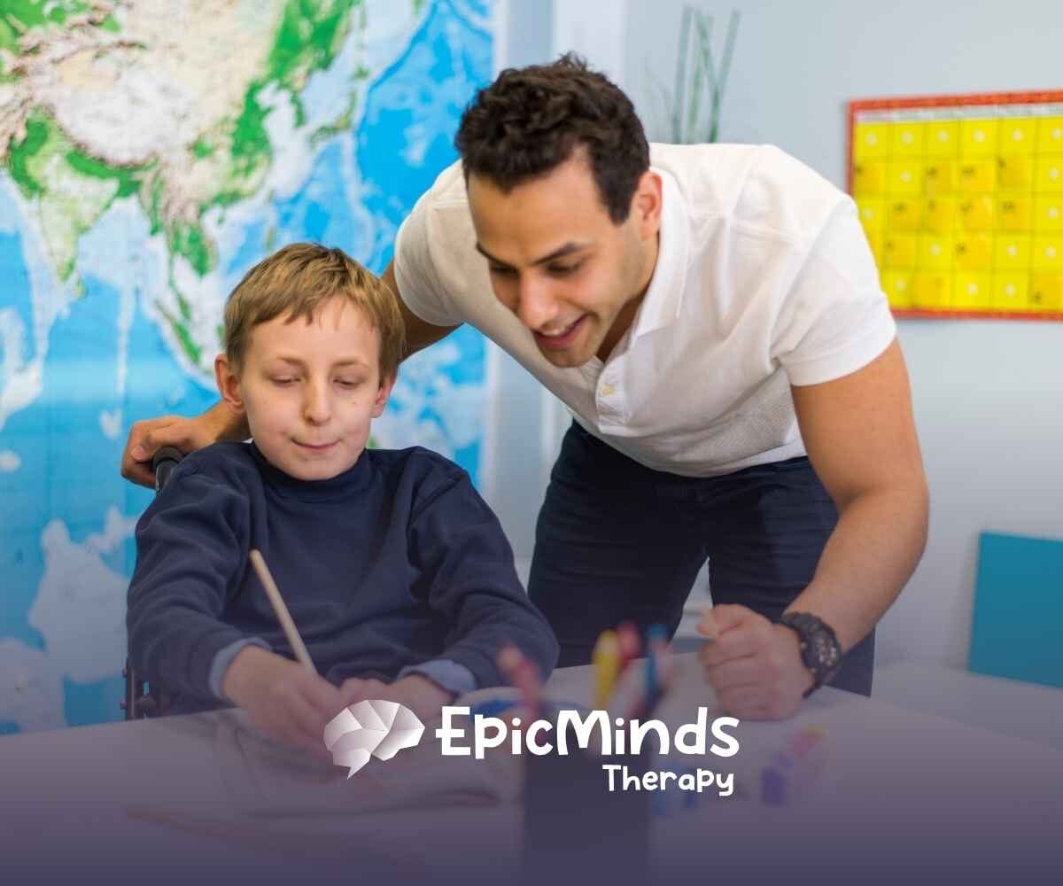 A smiling adult assists a child in a wheelchair with painting in a classroom setting. A world map and colorful calendar are visible in the background.