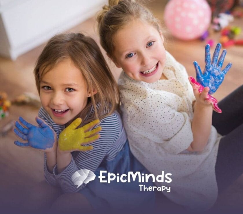 Two autistic girls sitting on the floor smiling and showing their hands covered in colorful paint during ABA therapy.