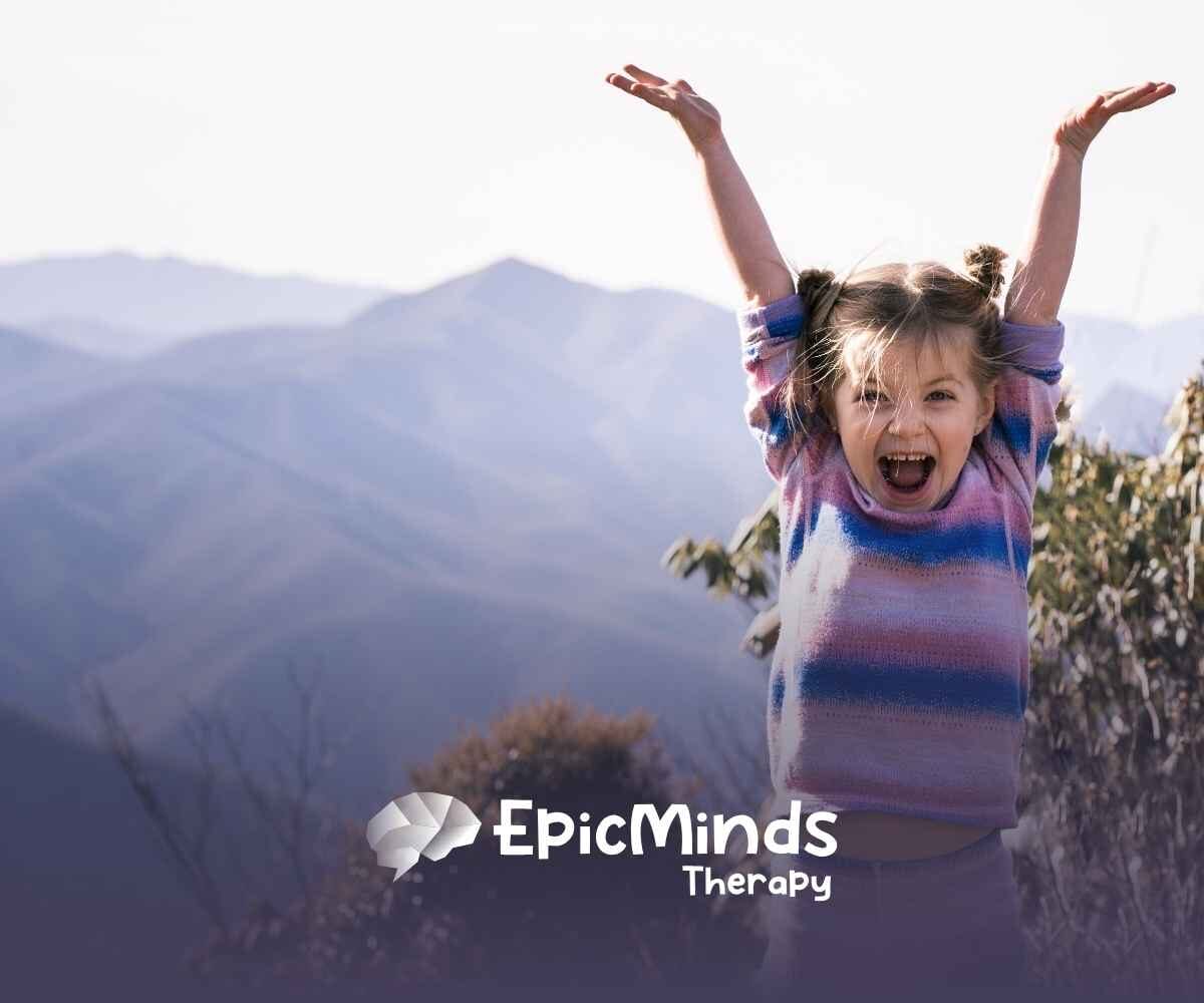 Young girl with pigtails, wearing a colorful sweater, joyfully raises her arms against a backdrop of misty mountains and foliage.