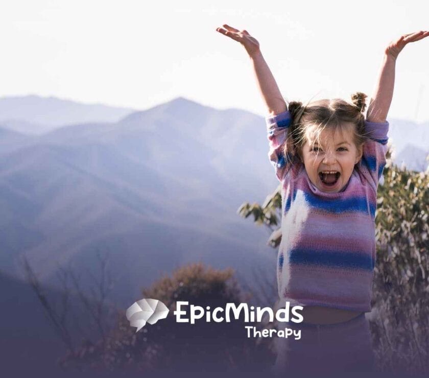 Young girl with pigtails, wearing a colorful sweater, joyfully raises her arms against a backdrop of misty mountains and foliage.