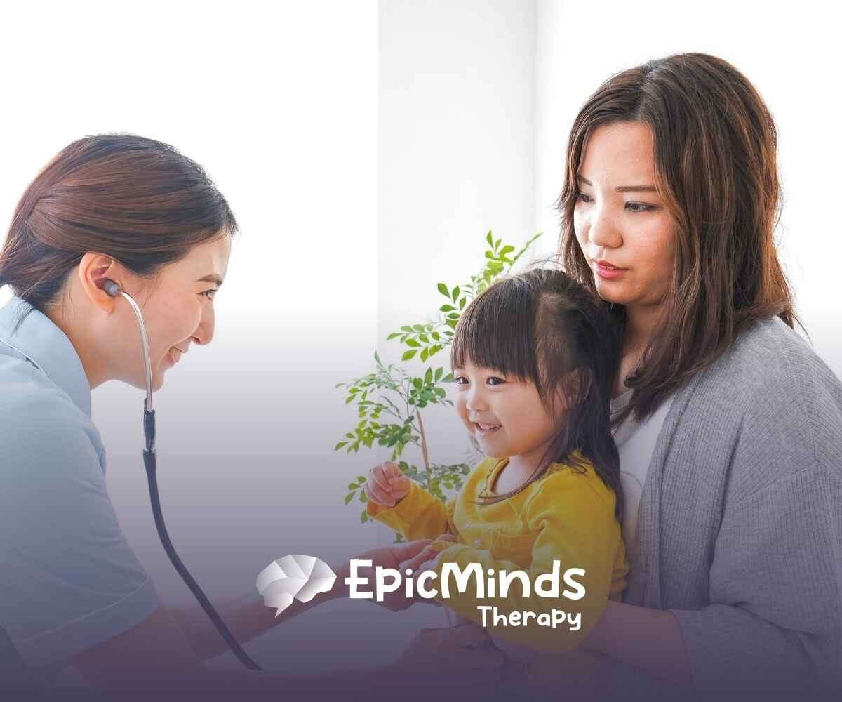 A nurse with a stethoscope smiles at a smiling toddler in a yellow shirt, held by a woman. The setting is bright and caring, with a small plant in the background.