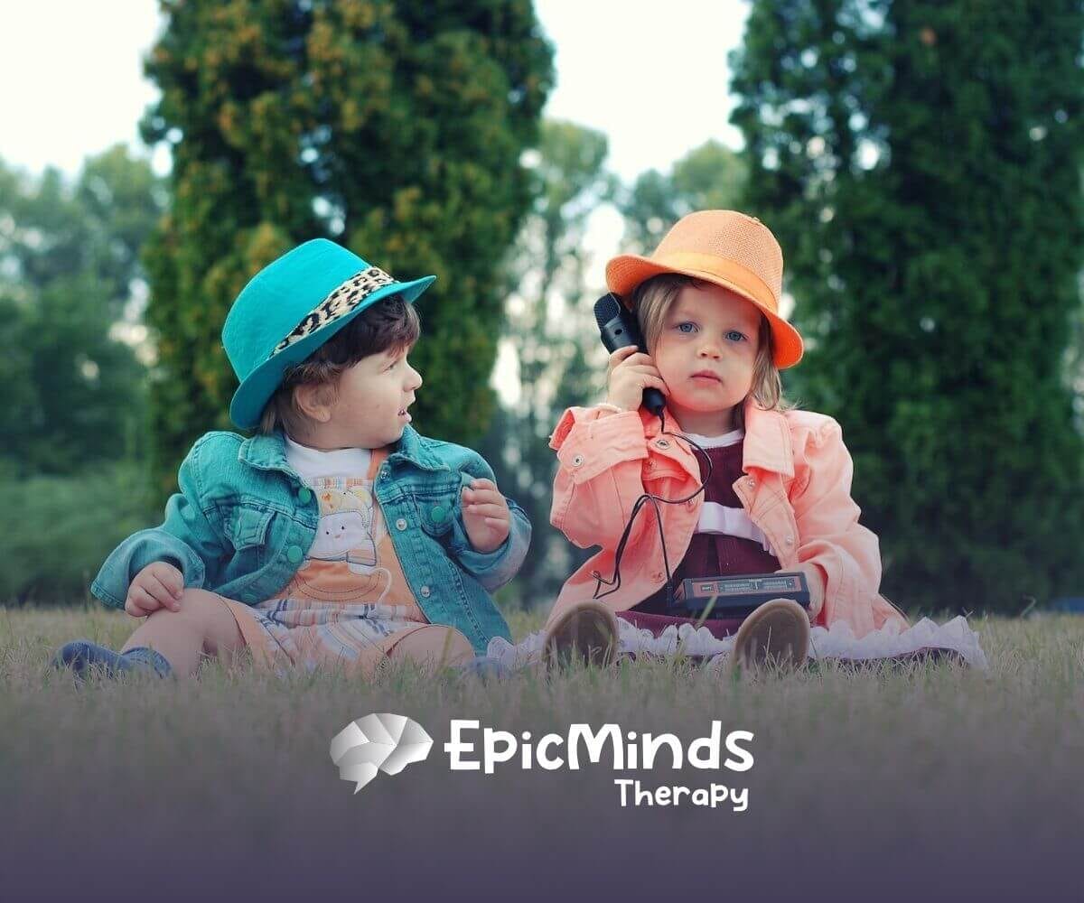 Two autistic toddlers in colorful hats sitting on grass with a toy phone and playing after their ABA therapy.