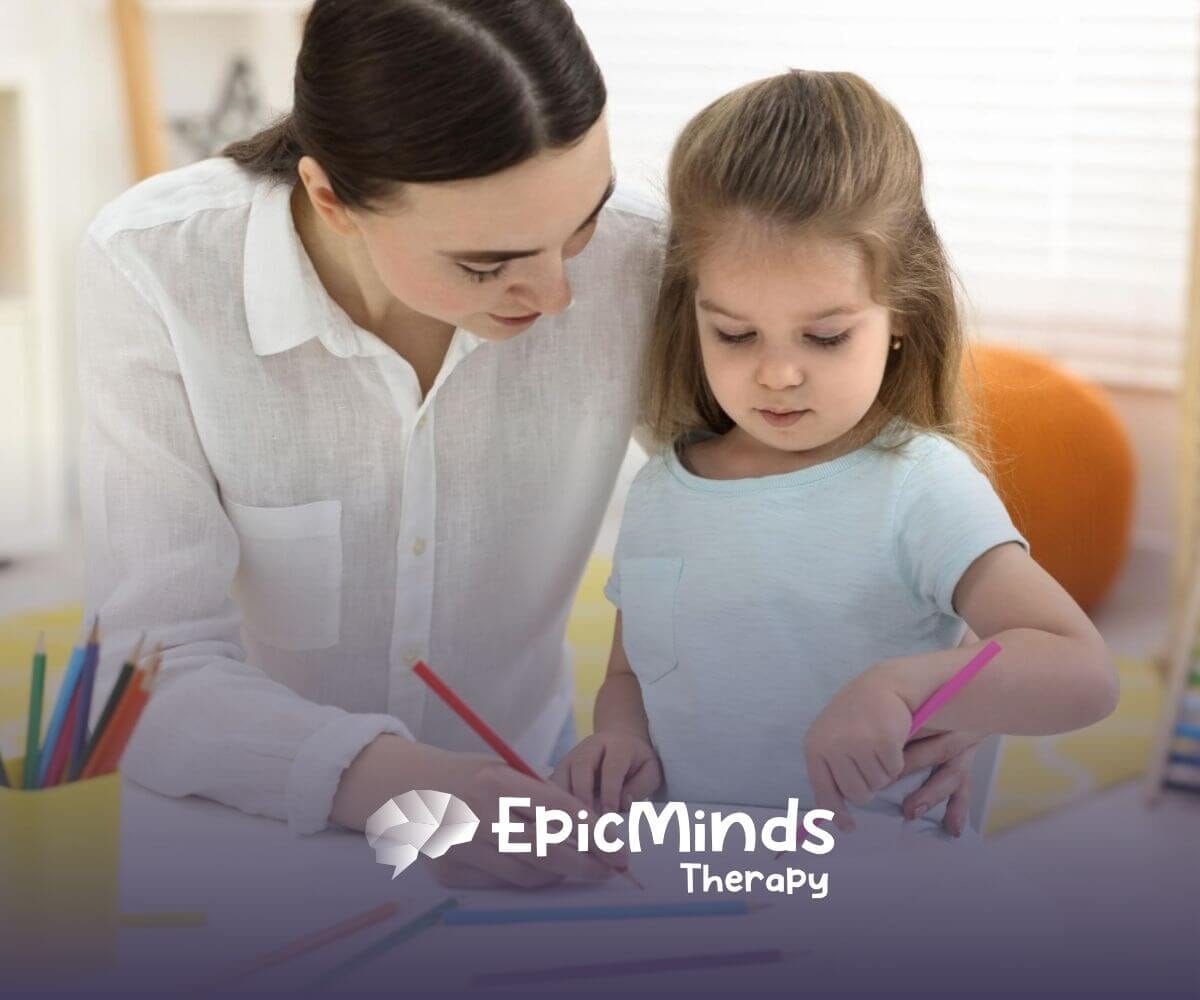 Therapist helping young girl draw with colored pencils at a table during ABA therapy.