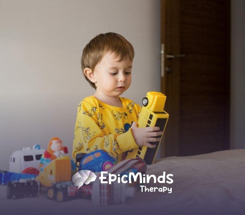 Toddler with autism holding a yellow toy bus while playing with toy vehicles.
